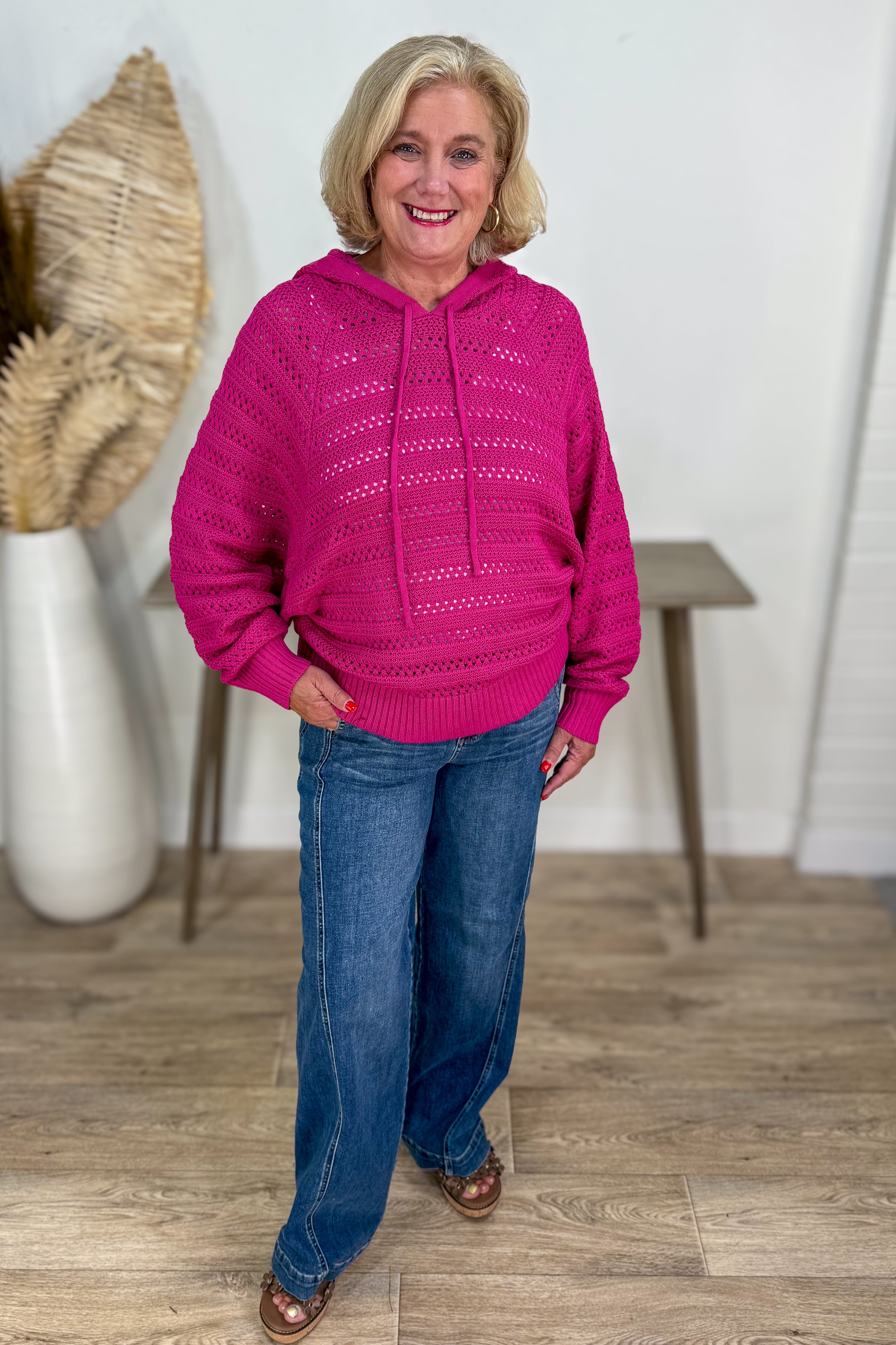 Woman wearing a pink hoodie and blue jeans standing indoors with a neutral background