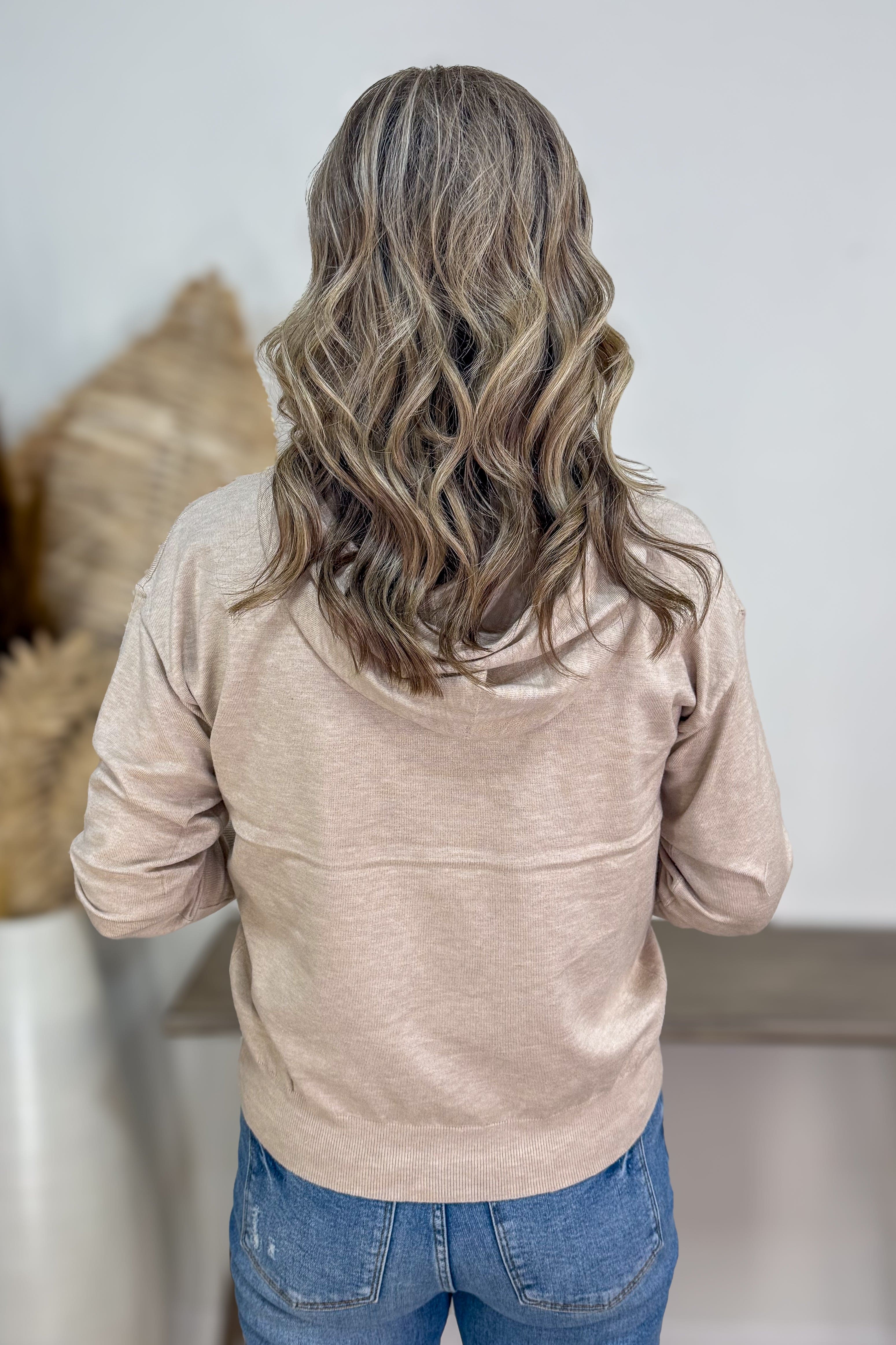 Person wearing a beige jacket and blue jeans with wavy hair, standing against a neutral background.