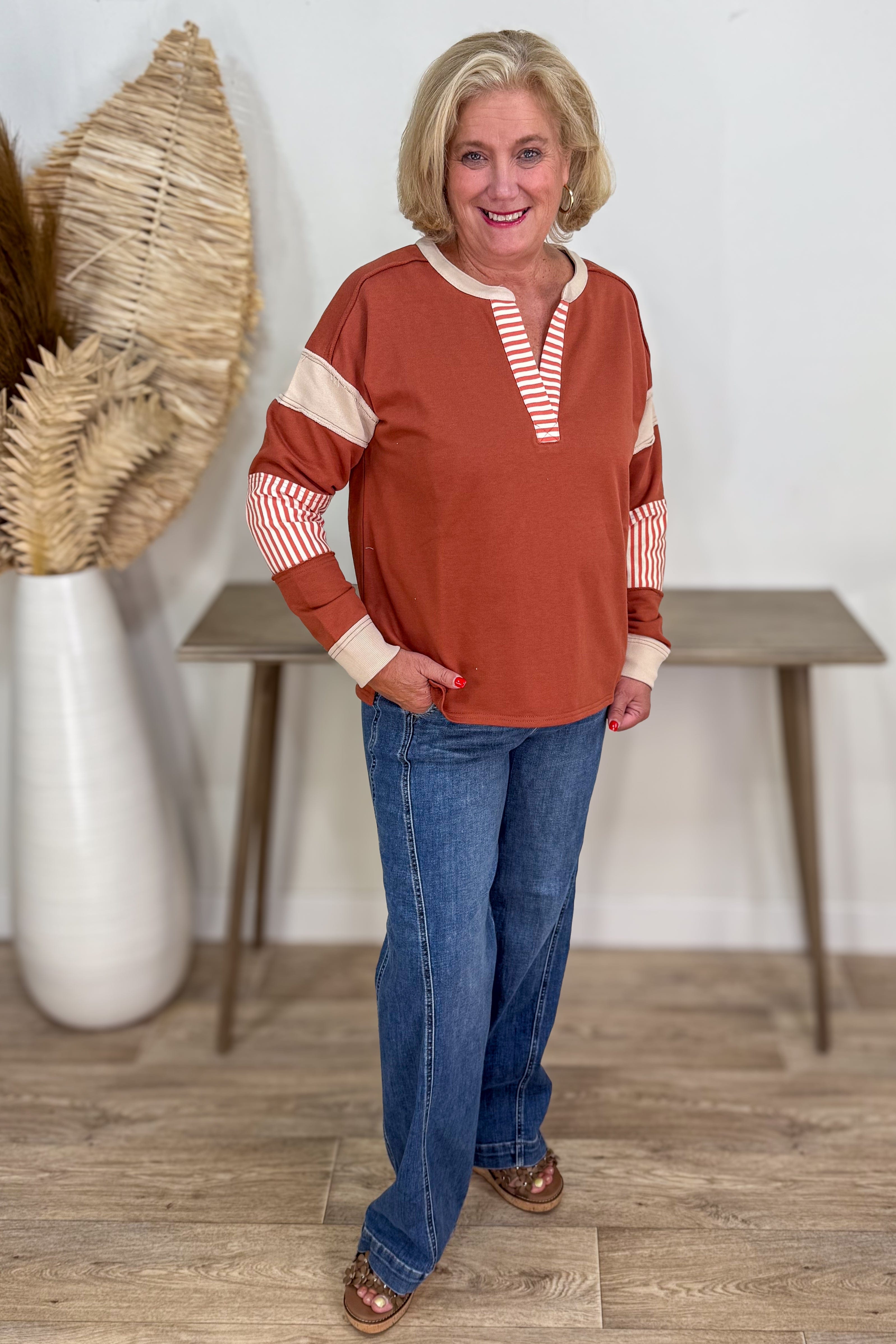 Woman wearing a rust-colored top with white stripes and blue jeans standing indoors.