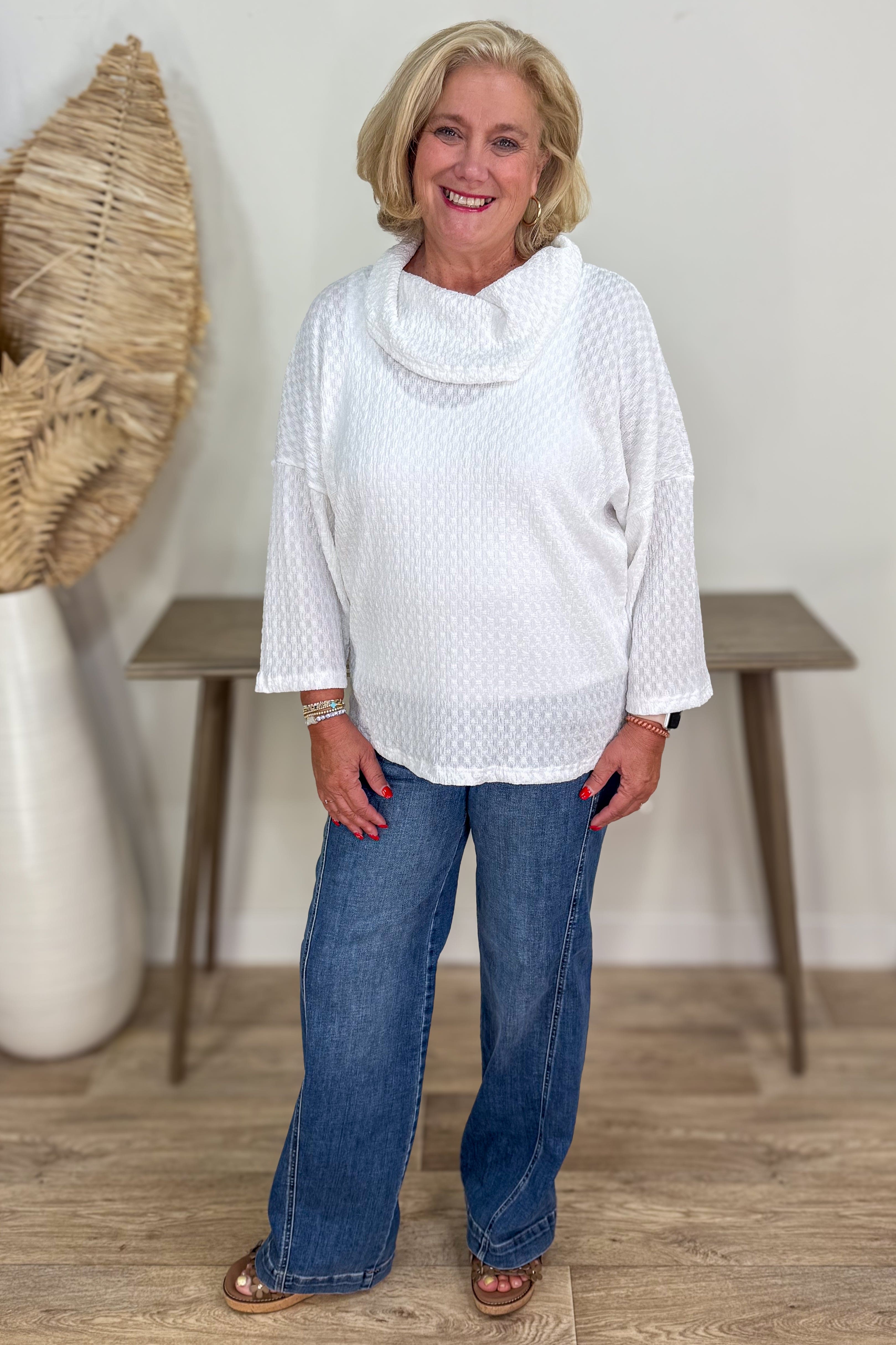 Woman wearing a white top and blue jeans standing in a room with a table and decorative items.