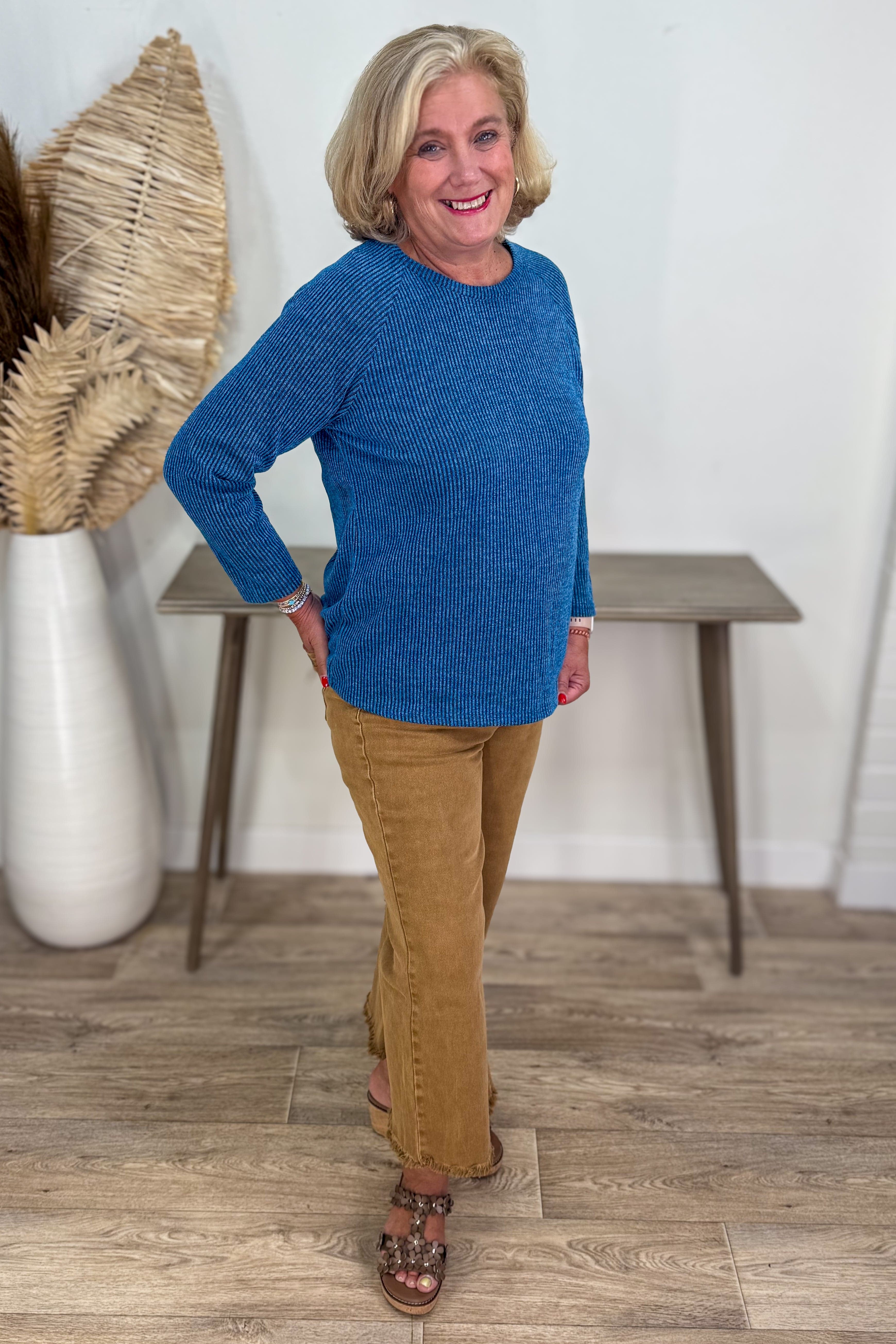 Woman wearing a blue sweater and tan pants standing in a room with a table and decorative items.