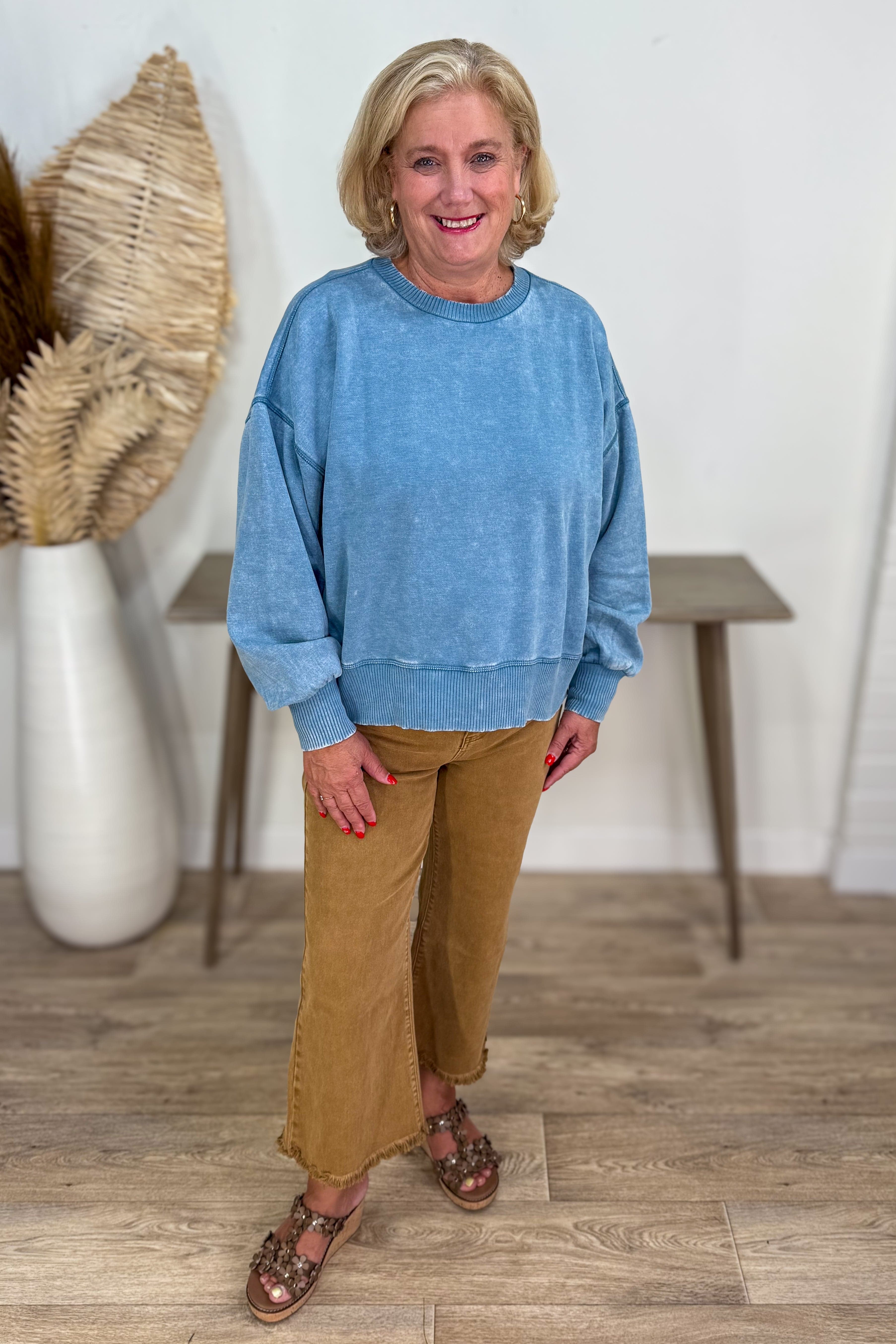 Woman wearing a blue sweater and brown pants standing in a room with wooden flooring and a vase.