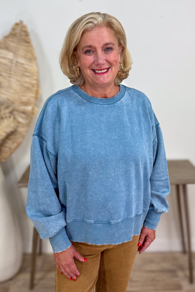 Woman wearing a blue sweater and brown pants indoors.