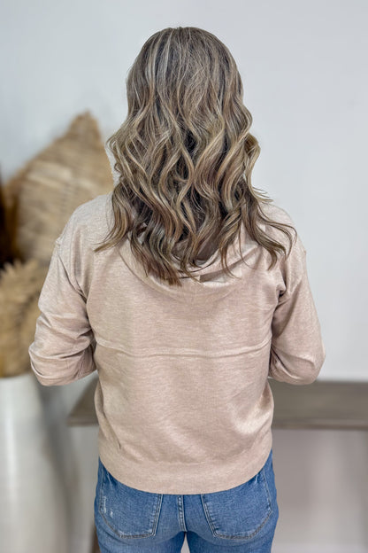 Person wearing a beige jacket and blue jeans with wavy hair, standing against a neutral background.