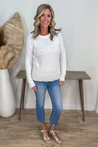 Woman wearing a white sweater and blue jeans standing in a room with a wooden floor and white wall.