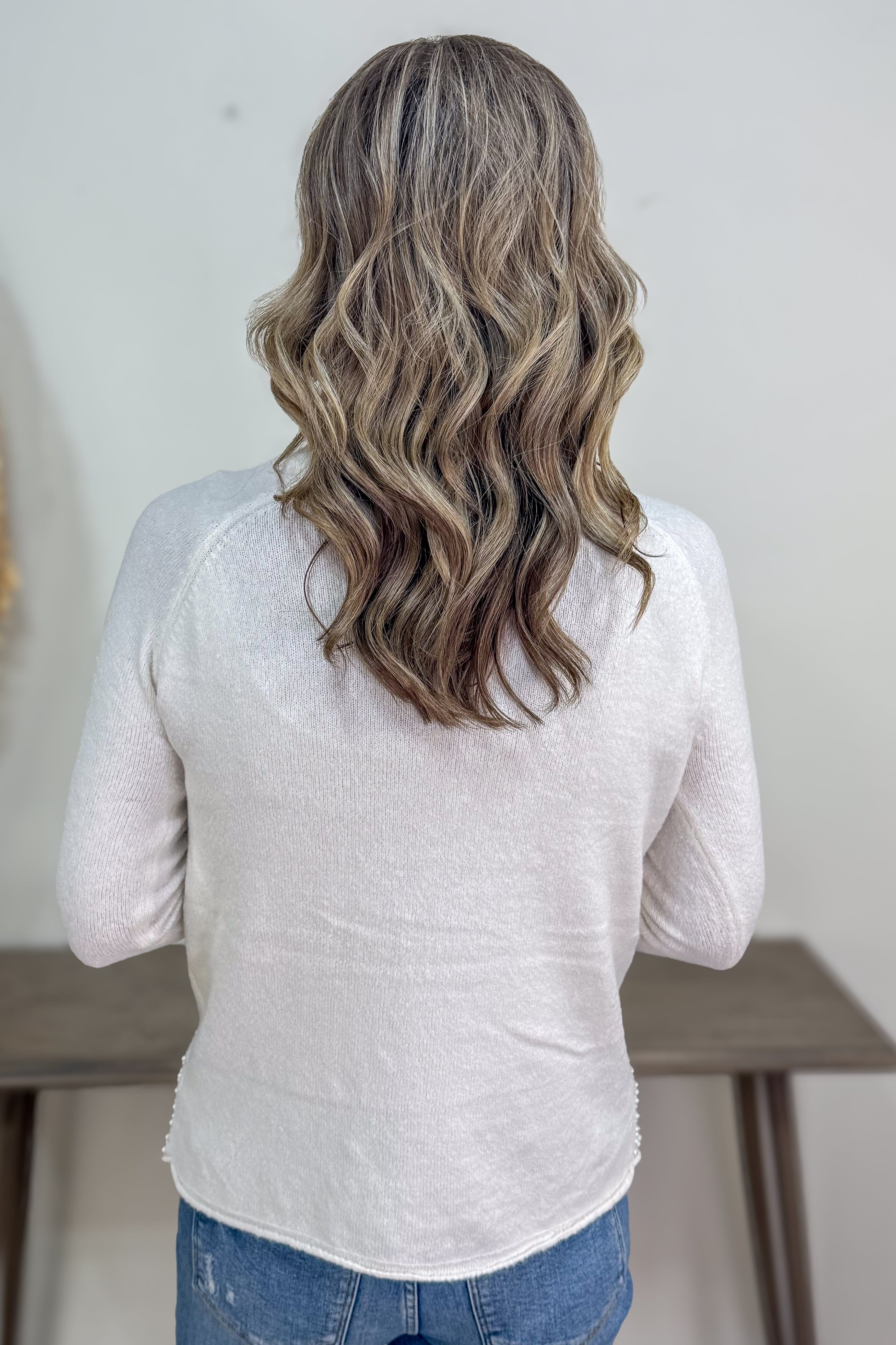 Person with wavy hair wearing a light gray long-sleeve shirt and blue jeans, standing against a neutral background.
