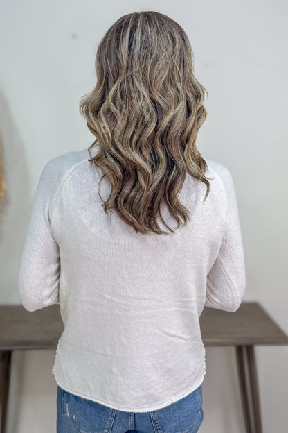 Person with wavy hair wearing a light gray long-sleeve shirt and blue jeans, standing against a neutral background.